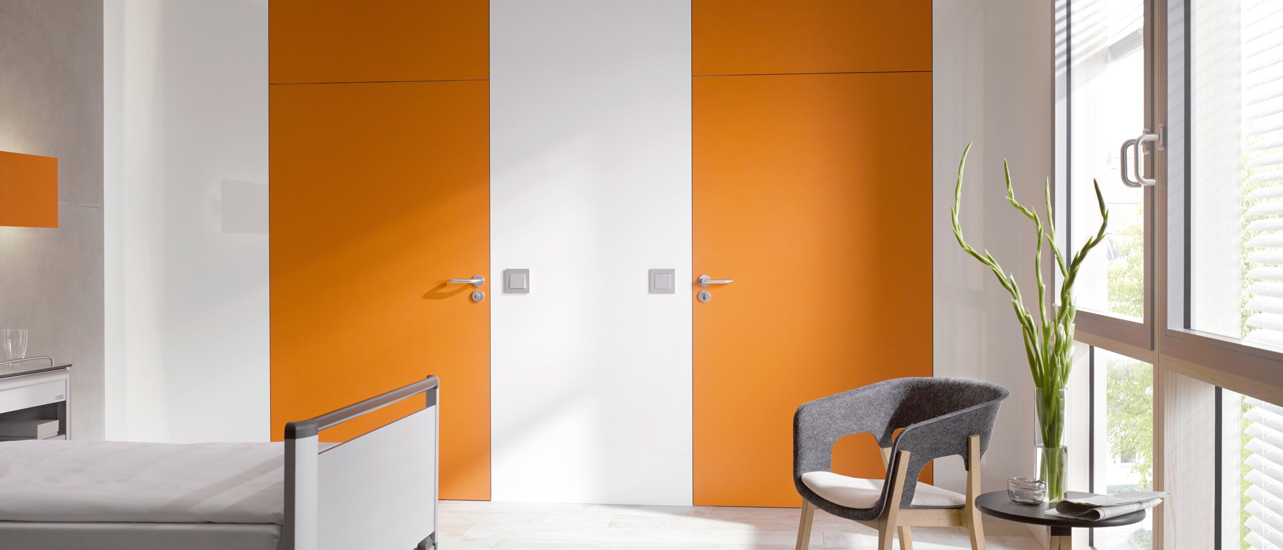 White hospital room with two orange doors fitted with a lever handle in the colour grey made of polyamide